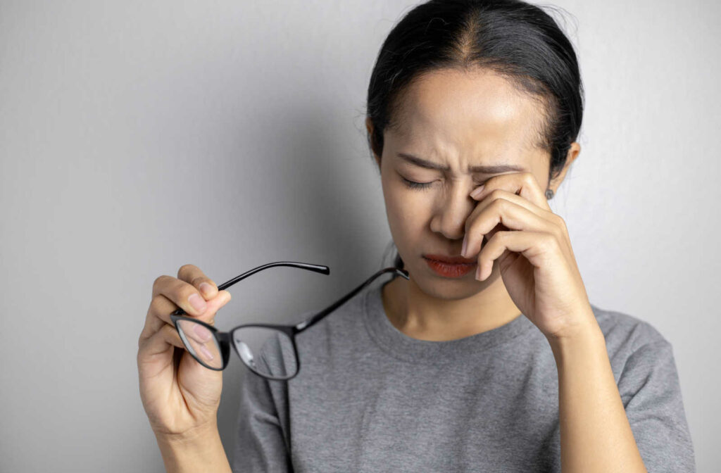 A young woman holding her glasses in her right hand as she rubs her dry eyes using her left hand.