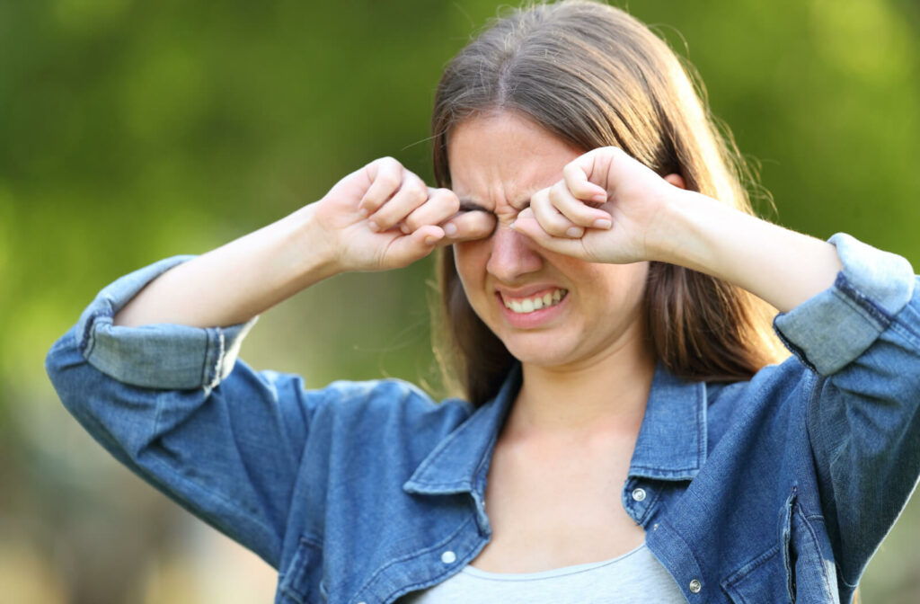 A woman outdoors is experiencing discomfort from dry and itchy eyes. Frustration is evident on her face as she scratches her eyes with both hands.